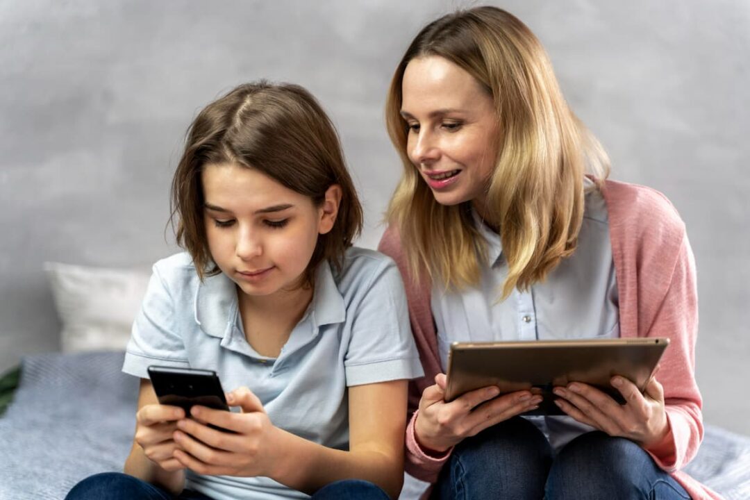 A imagem mostra uma mulher segurando um tablet e olhando a tela do celular da criança ao seu lado.