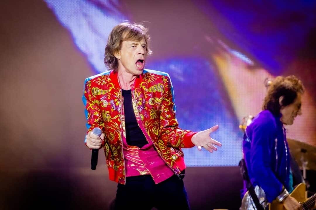 Imagem de Mick Jagger, vestido com roupas vermelhas, segurando um microfone em cima do palco. Ele também é pai do filho de Luciana Gimenez.