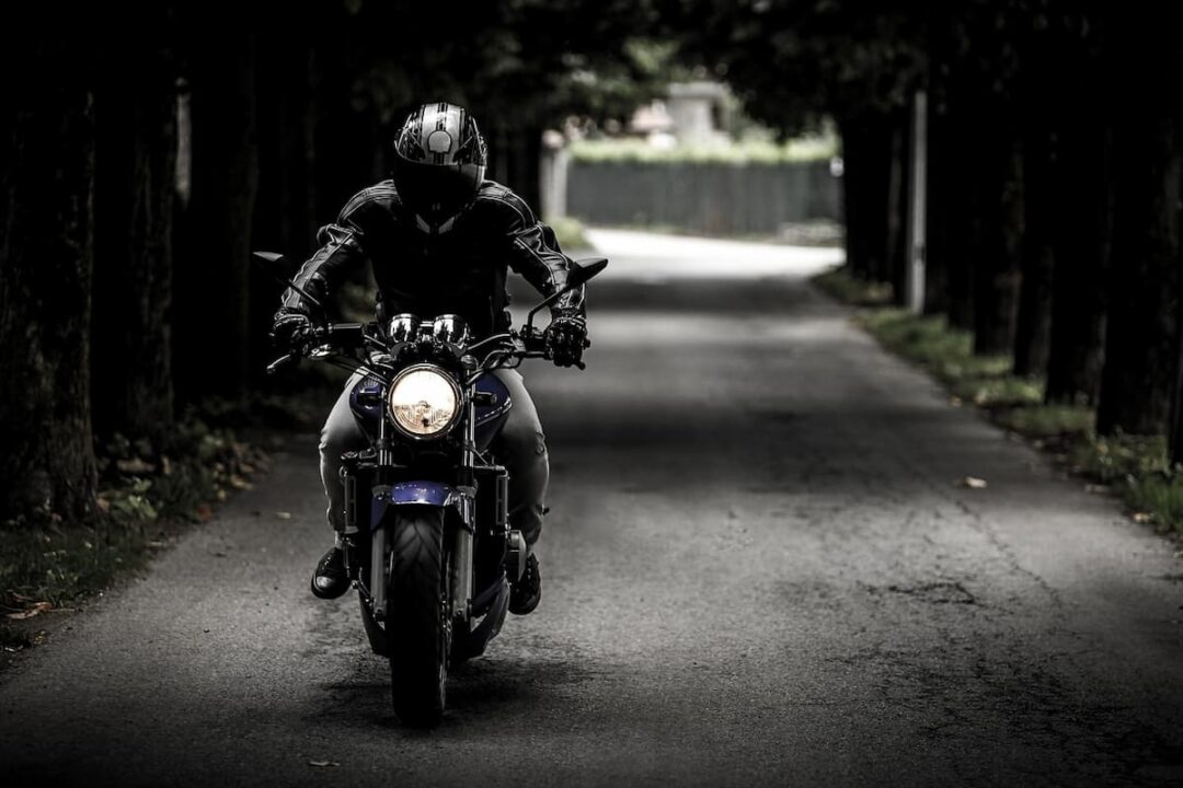 A imagem mostra um homem, com casaco e capacete preto, em uma moto.