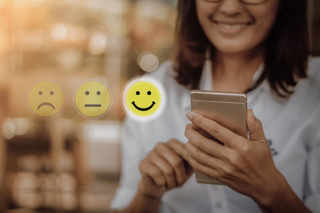 mulher sorrindo com o celular na mão feliz com o atendimento. Emojis com cara de feliz, neutro e triste