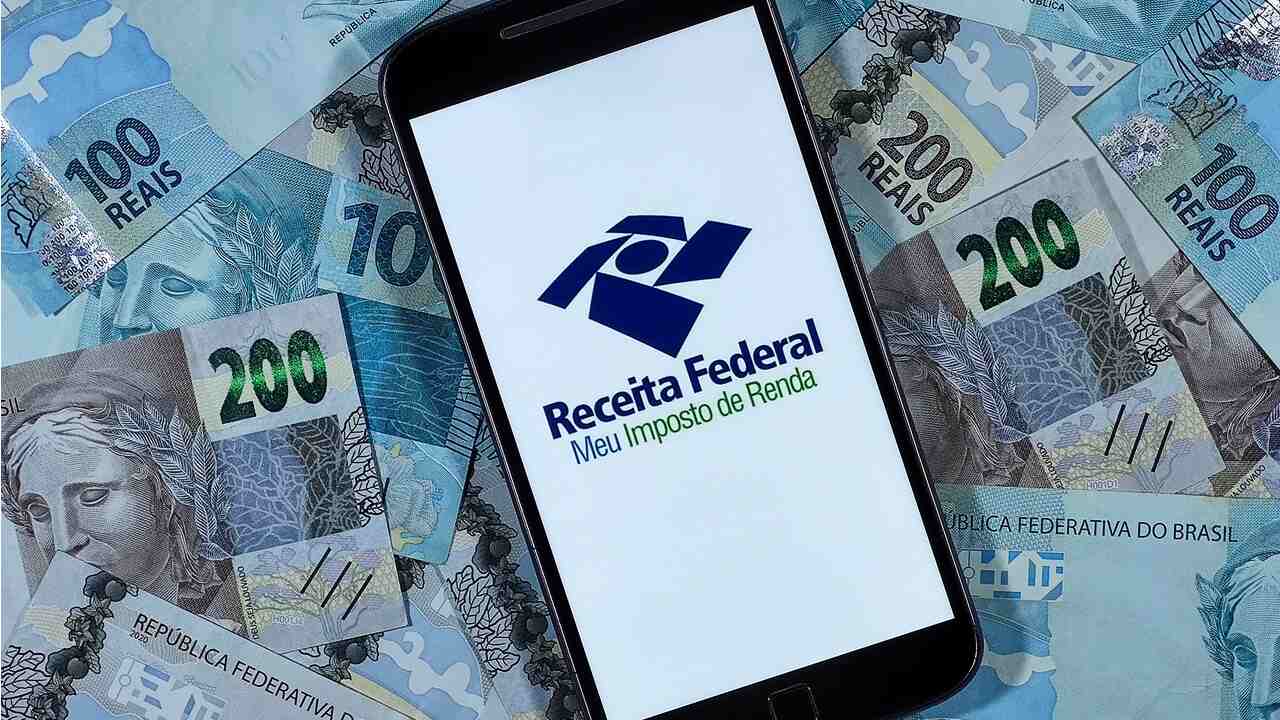 No centro de notas de 100 e 200 reais, celular com logo da Receita representando a restituição do IR.