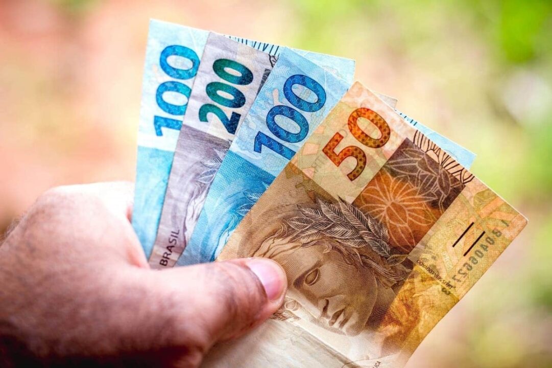 Mão segurando notas de R$50, R$100 e R$200 representando auxílios do governo