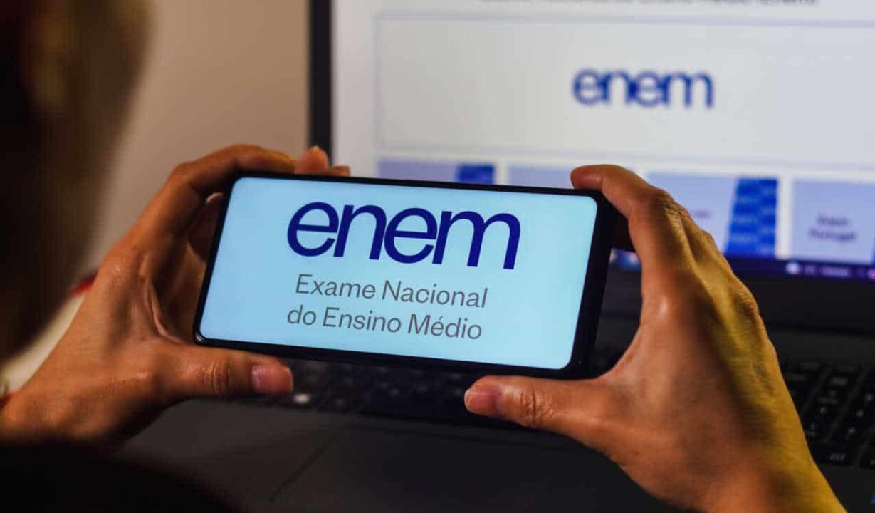 Pessoa segurando um celular com Enem escrito na tela. No fundo um computador com o site do Enem aberto