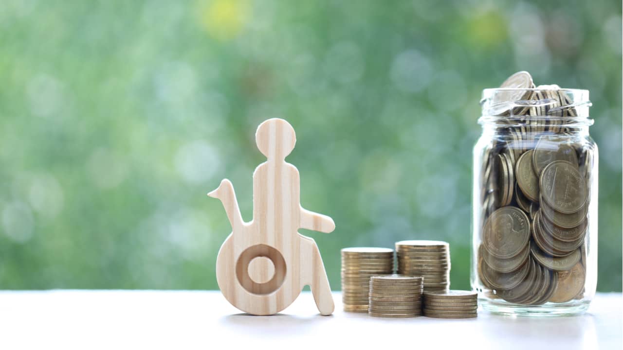 Miniatura de pessoa em cadeira de rodas ao lado de pilhas de moedas e pote de moedas