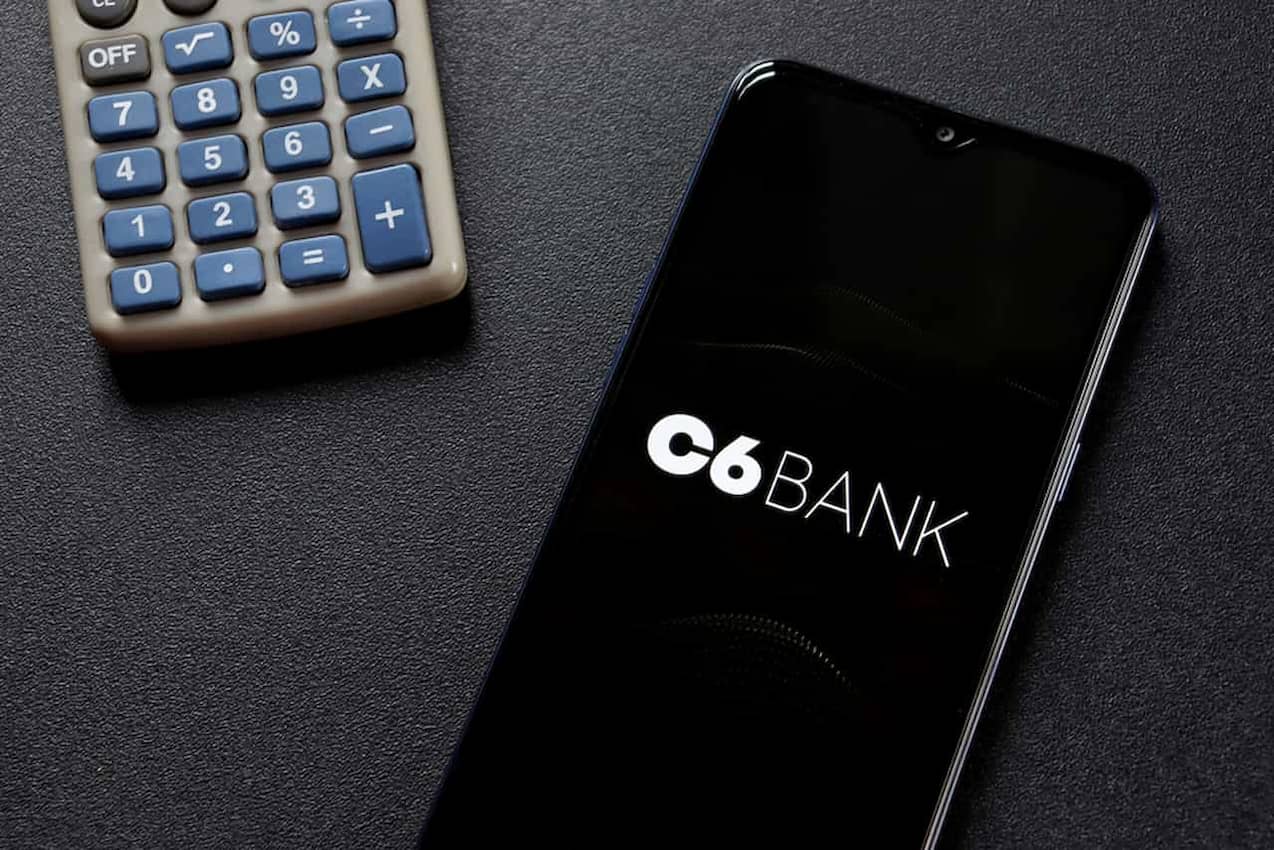 Ao lado de uma calculadora, celular exibe a logomarca do C6 Bank em fundo preto