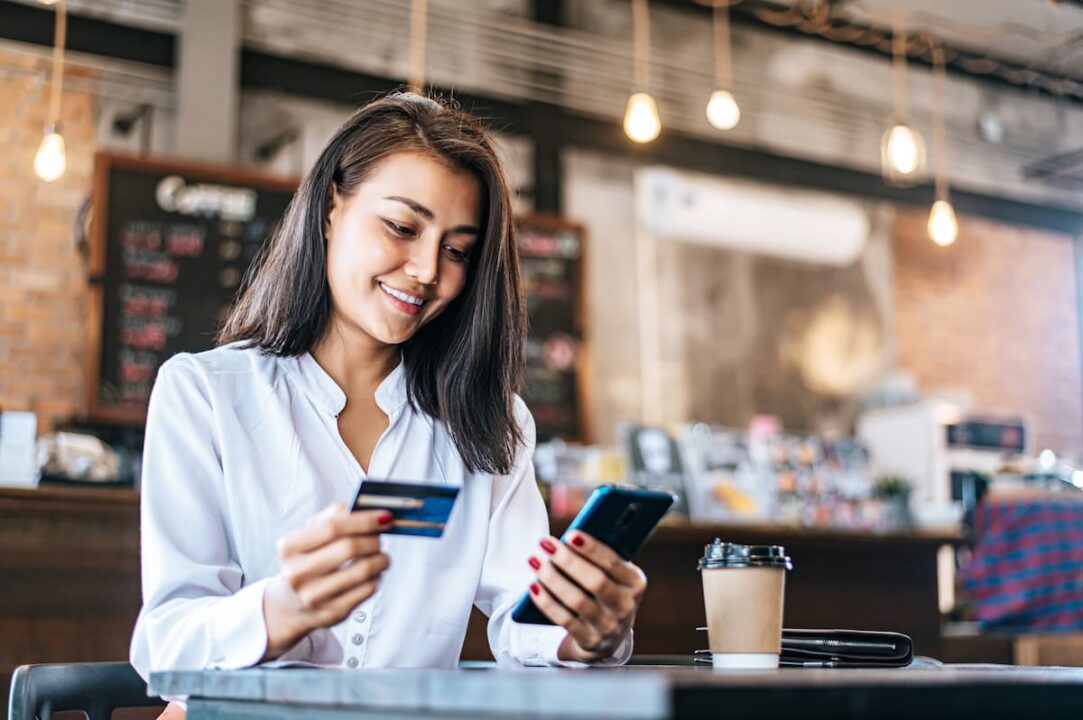 A imagem mostra uma mulher segurando um cartão e olhando a tela do celular.