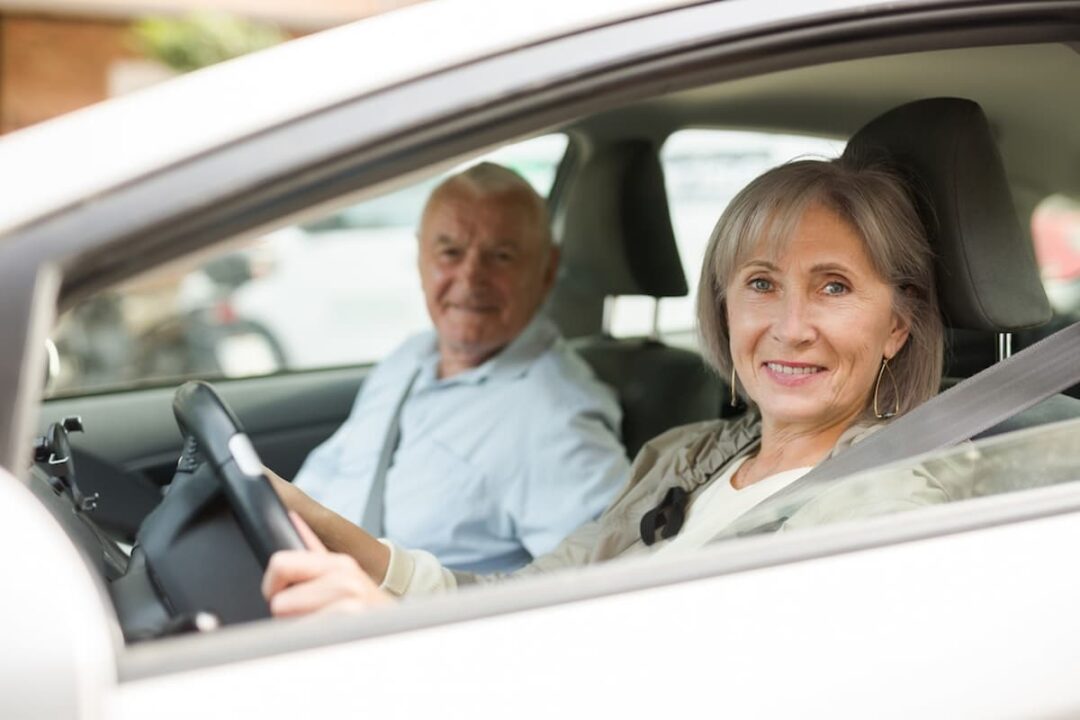 Casal de idosos em um carro. Mulher com as mãos no volante e sorrindo.