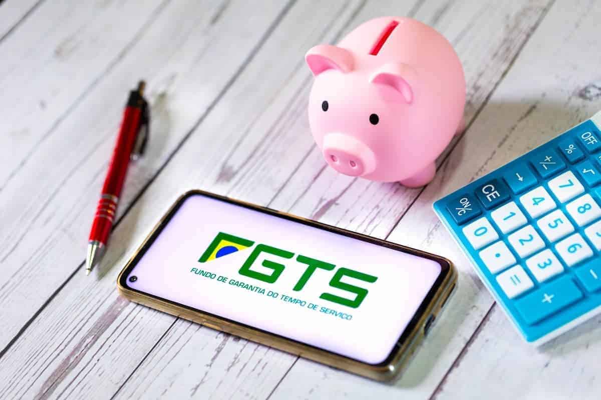 A imagem mostra um celular com a logo do app do FGTS, uma caneta, calculadora e um cofrinho para moedas em forma de porco.