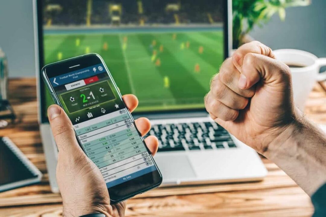Computador passando futebol na tela, uma mão segurando celular com aplicativo de aposta e outra mão cerrada comemorando resultado positivo depois de apostar no time vencedor