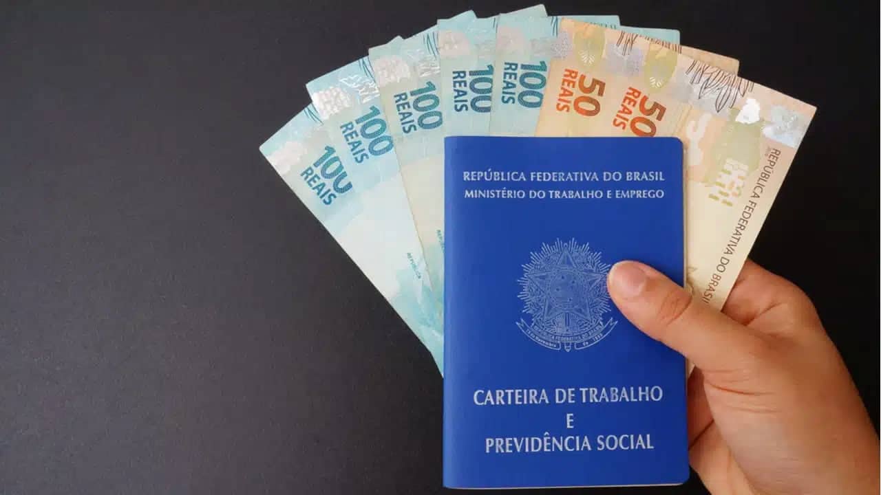 Imagem de mão segurando uma carteira de trabalho, com notas de 50 e 100 reais dentro dela, representando reajuste do salário  em 2024mínimo.