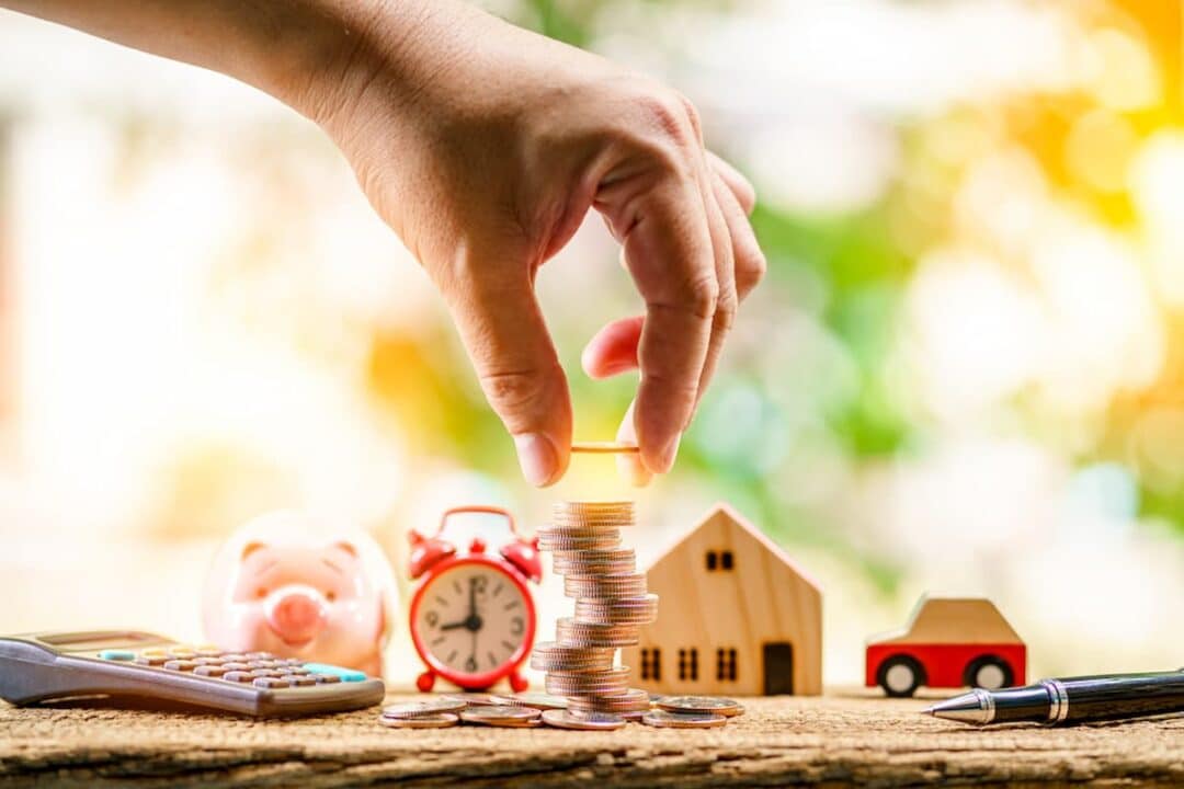 Imagem de mão colocando moedas, ao lado de porquinho, relógio, casa e automóvel, representando IPTU e IPVA