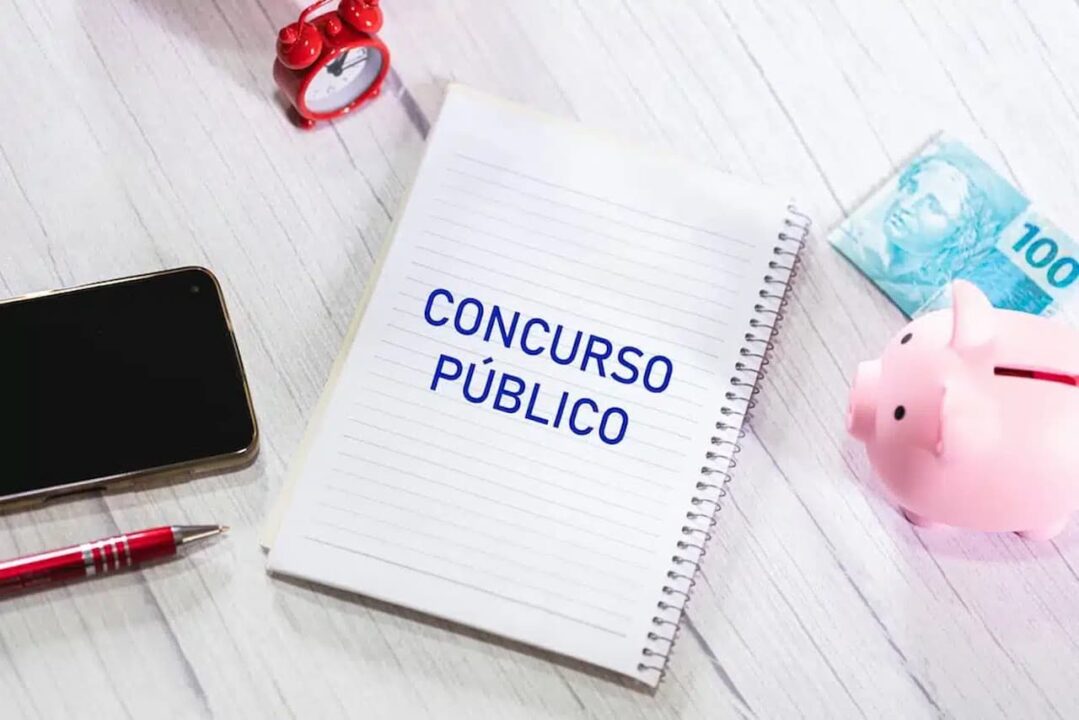 Caderno escrito "concurso público" e ao redor um cofrinho é formato de porco, nota de 100 reais, relógio e afins.
