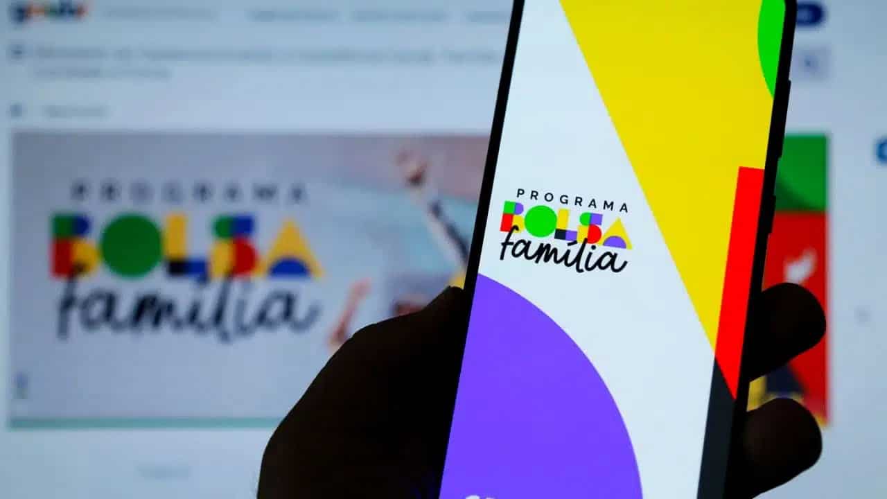 Uma mão segurando um celular com o aplicativo do Bolsa Família aberto. Ao fundo, a tela de um computador com o site para verificar aumento do Bolsa Família.