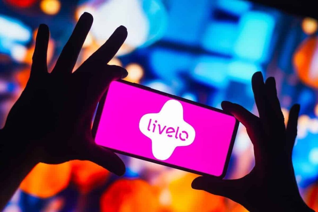 Mãos segurando celular com o logo da Livelo na tela, em um fundo desfocado colorido.