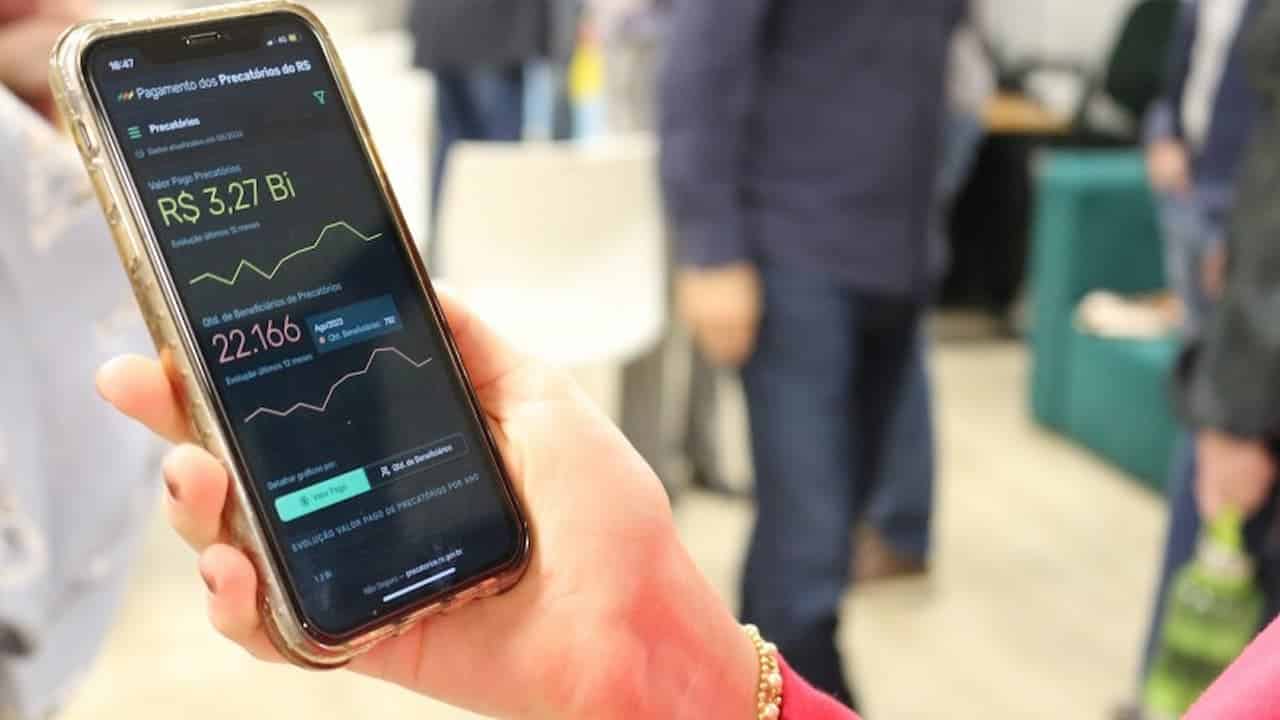 Mão segurando um celular com o Portal de Pagamentos dos Precatórios aberto.
