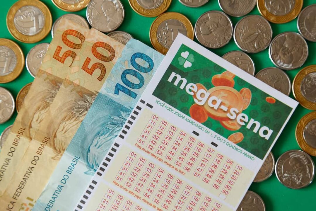 Cartela da Mega-Sena ao lado de notas de 50 e 100 reais em cima de moedas de 50 centavos e 1 real.