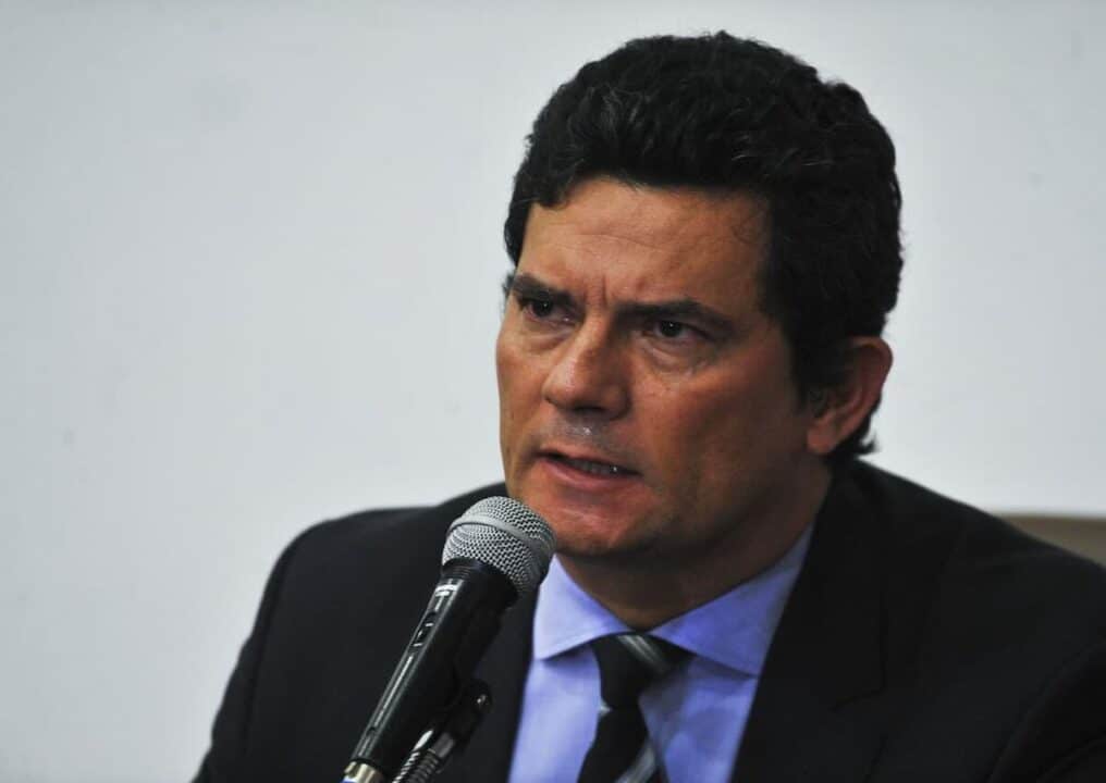 O ex-ministro da Justiça, Sergio Moro, discursando no microfone, com um fundo branco atrás.