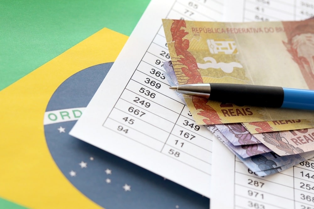 Imagem de caneta sobre notas de dinheiro, papĂ©is e bandeira do Brasil, representando o novo salário mĂnimo.