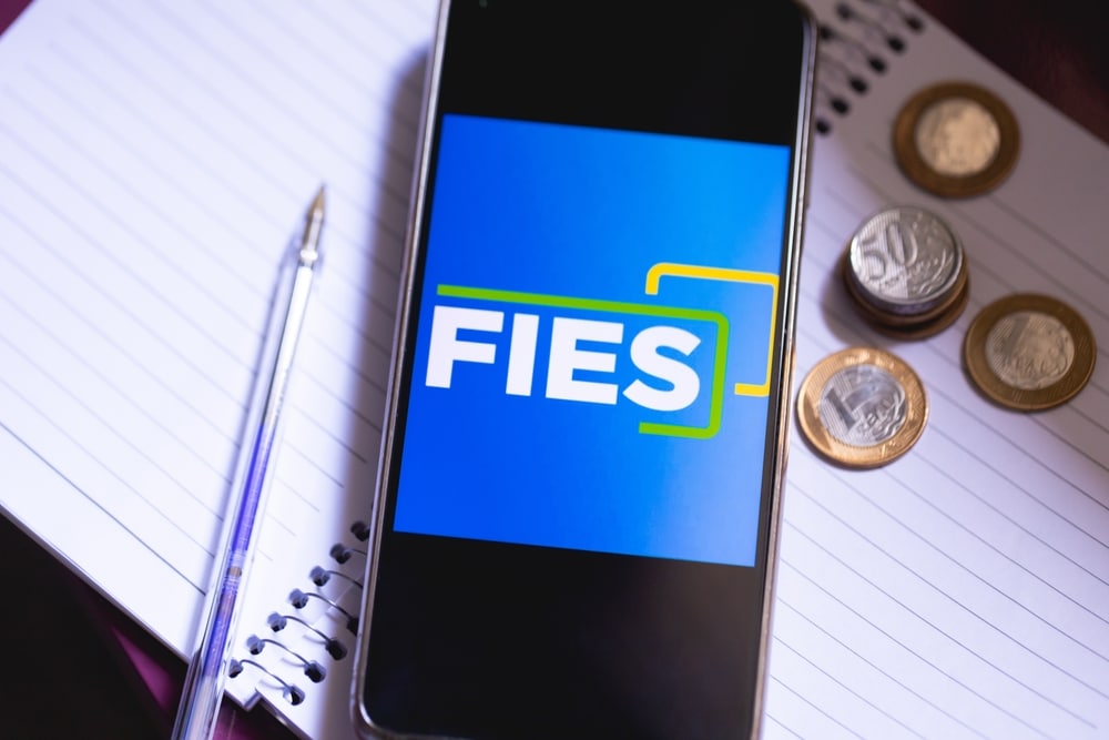 Celular com o nome "FIES" na tela, apoiado em um caderno, com algumas moedas do lado