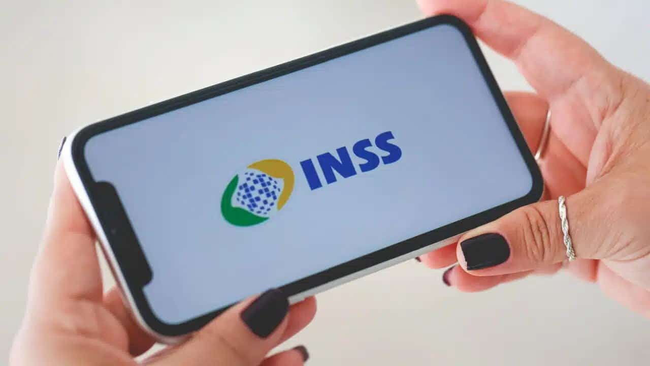 Mão segurando celular que mostra logo do INSS .