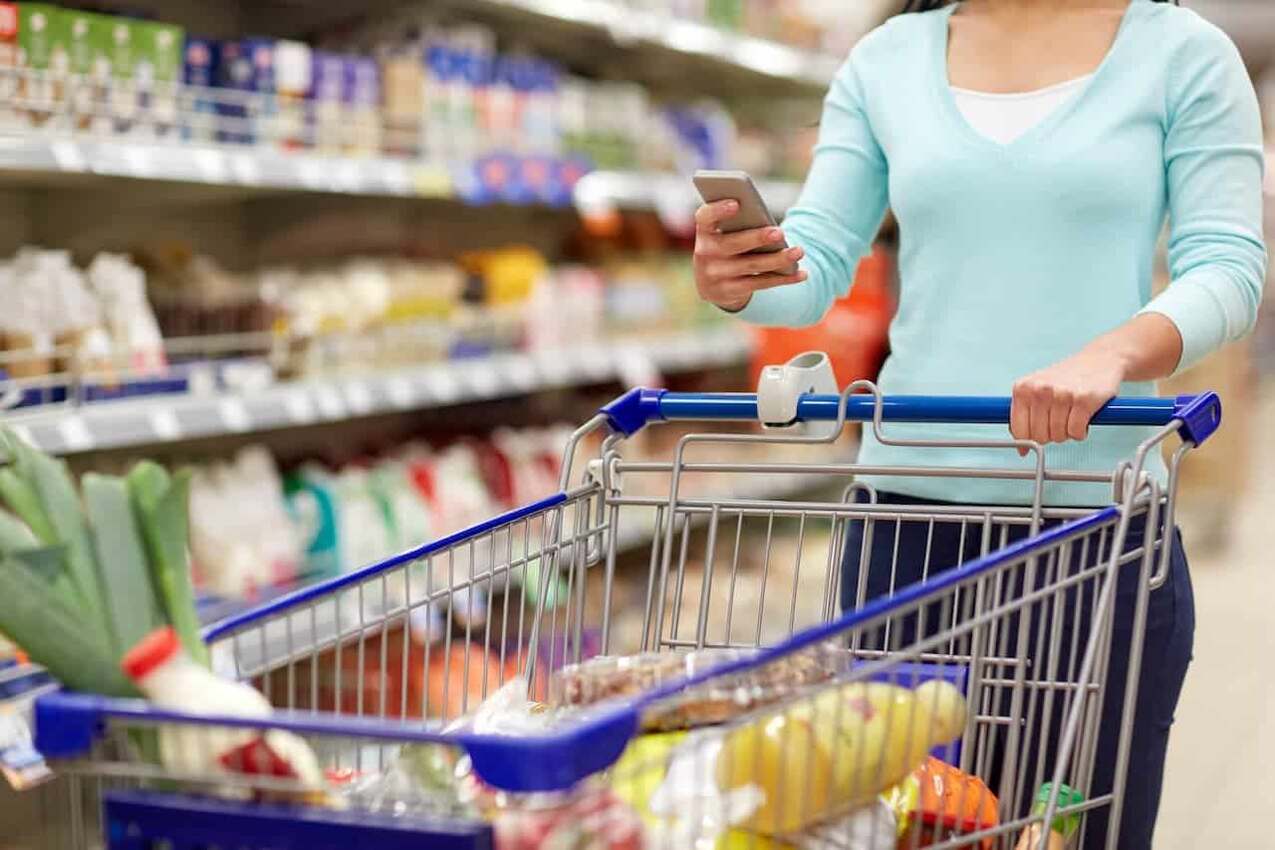 Mulher em rede de supermercado com carrinho de compras e segurando celular.
