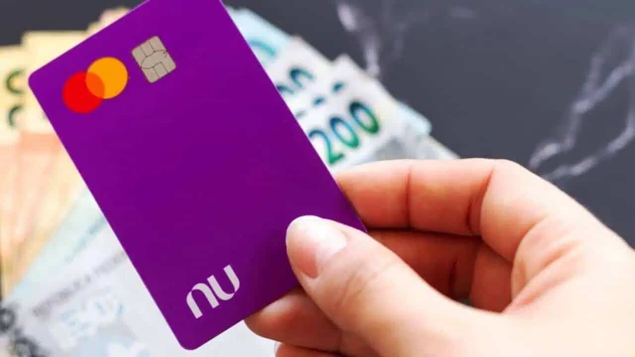 Mão segurando cartão do Nubank e cédulas de dinheiro ao fundo.