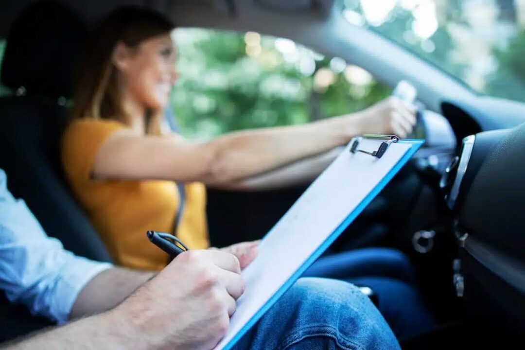 Mulher dirigindo carro enquanto homem segura uma prancheta avaliando e supervisionando o teste prático de direção.
