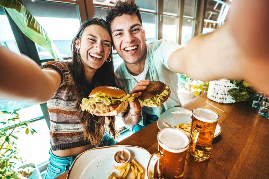 Casal jovem olhando para câmera comendo os melhores hambúrgueres em uma hamburgueria.