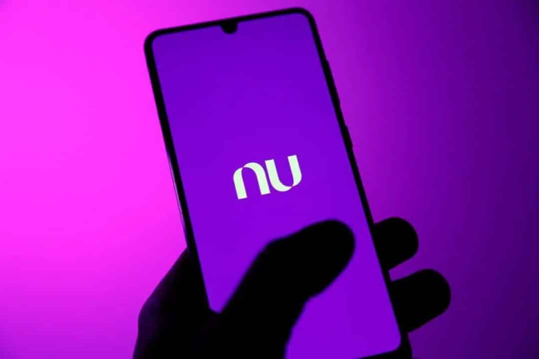 Mão segurando celular com logo do Nubank na tela.