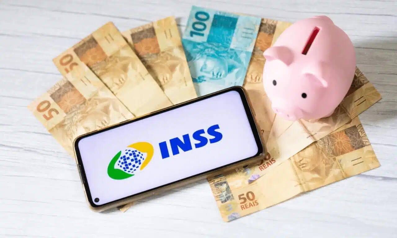 Notas de dinheiro com um cofrinho e o aplicativo do INSS aberto em um celular
