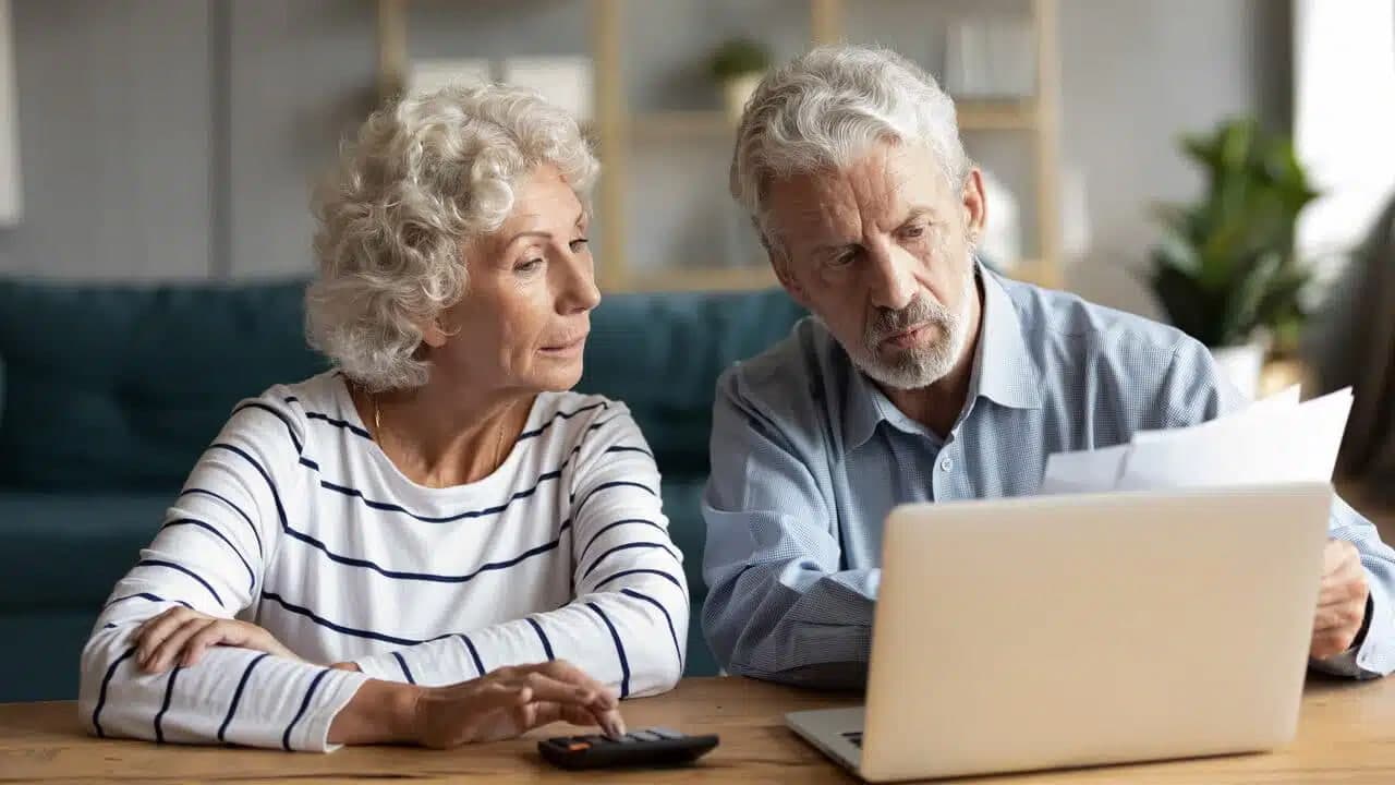 Mulher idosa e homem idosos sentados lado a lado e olhando para a tela de um notebook.
