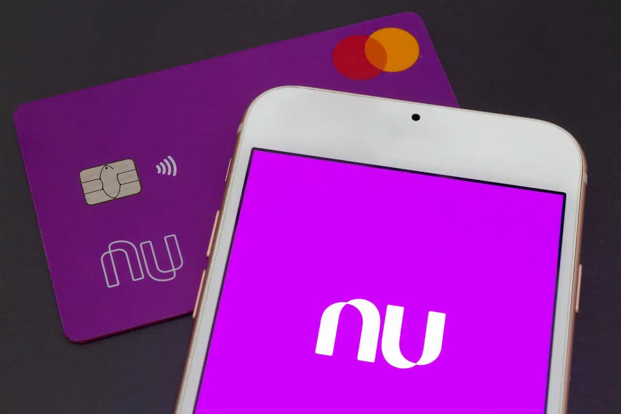 Imagem de um cartão de crédito do Nubank com logotipo antigo. Em cima de parte do cartão aparece um celular branco mostrando o logotipo atual do Nubank na tela