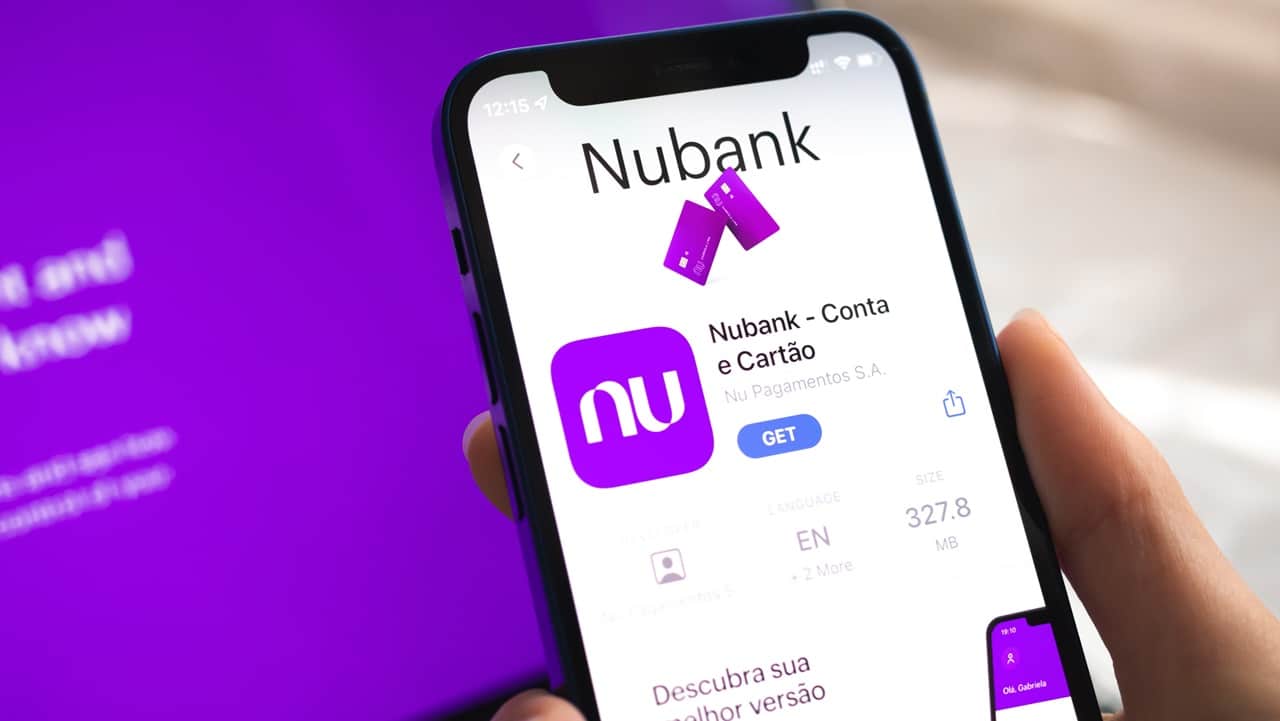 Imagem mostra a tela de um celular mostrando o aplicativo do Nubank para baixar. Ao fundo um notebook com tela de fundo roxo no site do Nubank. Nubank é um dos melhores bancos para guardar dinheiro