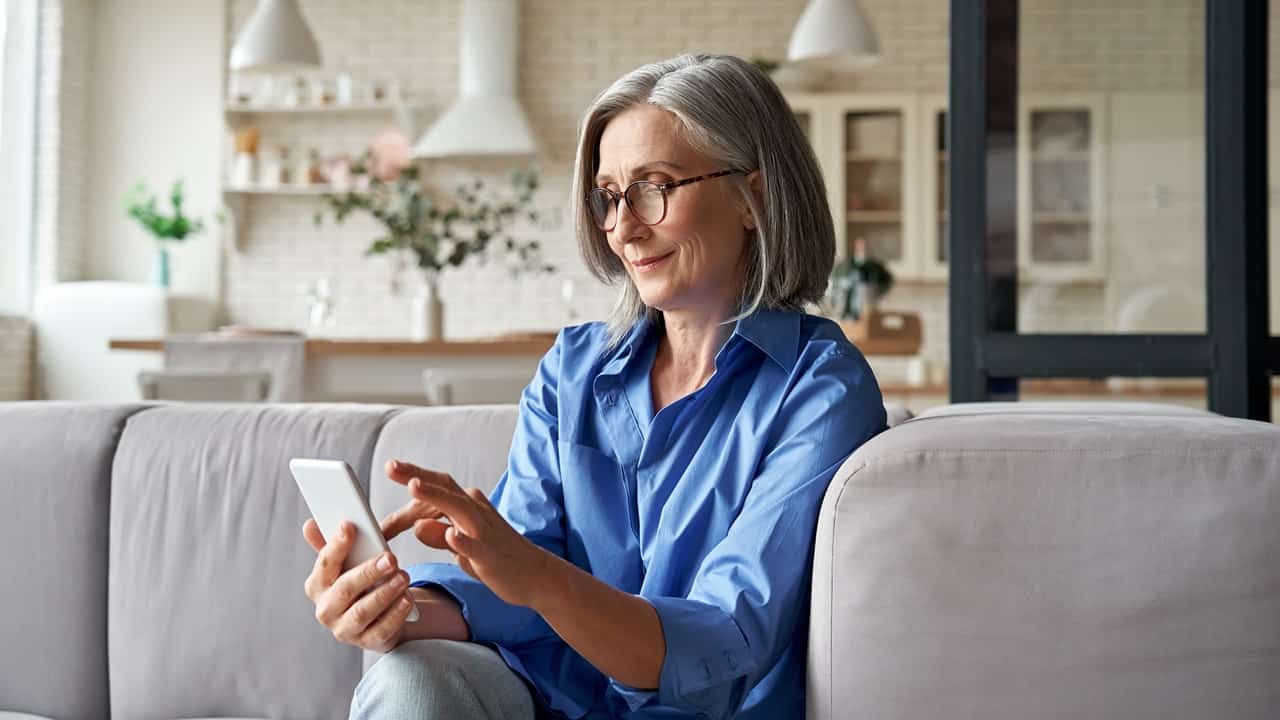Imagem mostra mulher idosa magra com cavelos grisalhos com um celular na mão baixando documentos digitais. Ela está sentada em um sofá cinza em uma sala.