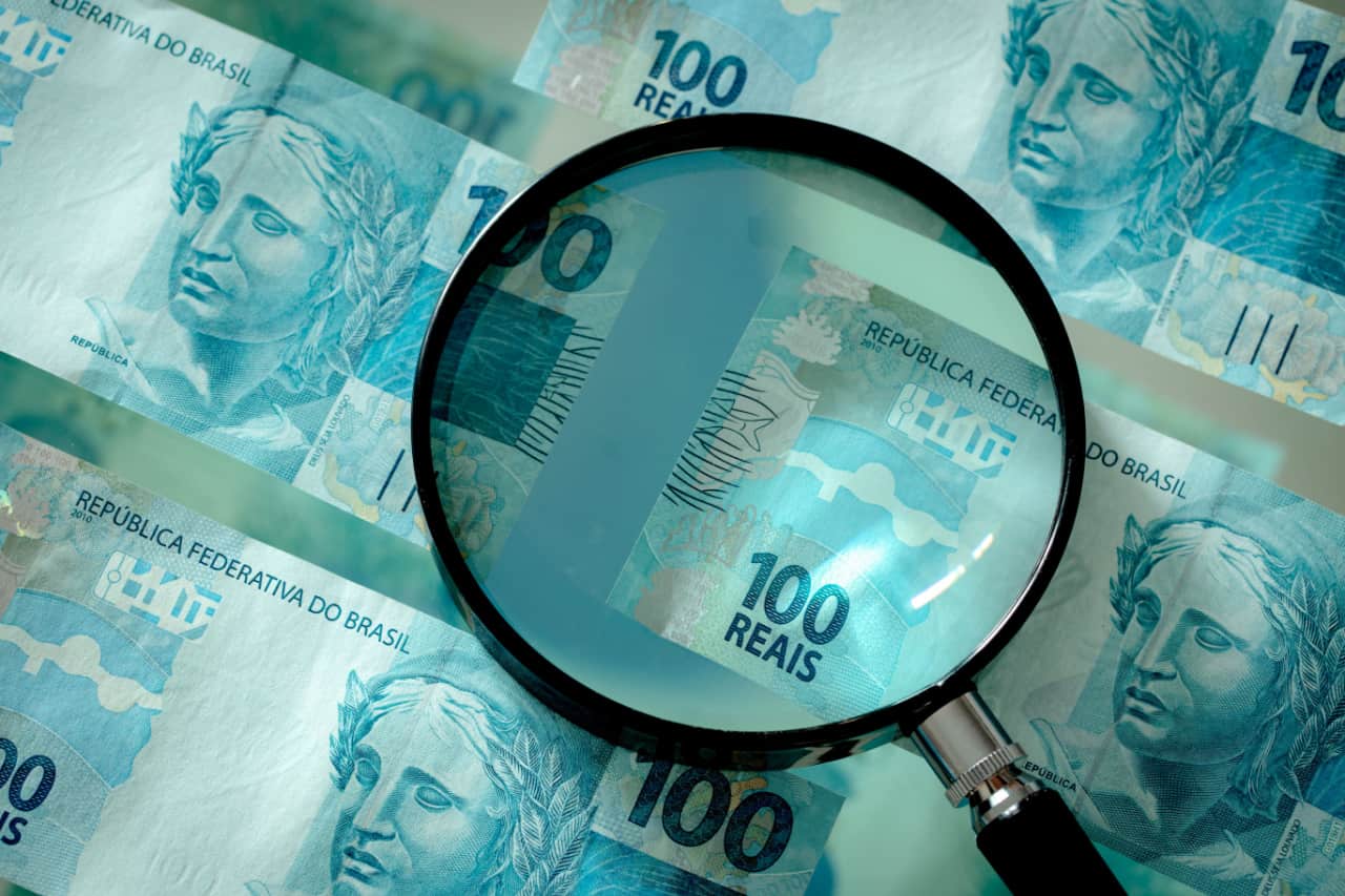 Notas de dinheiro brasileiro de 100 reais sob uma lupa com lente de aumento.