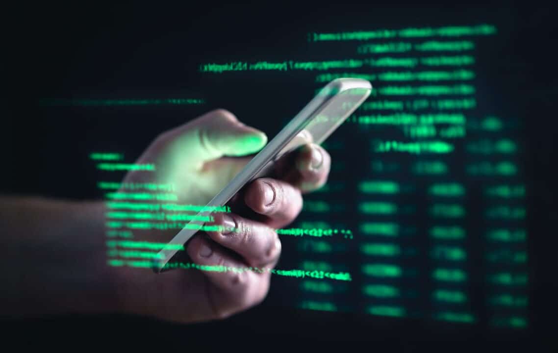 Imagem de uma mão de pessoa branca pegando um celular cinza claro, simbolizando um celular clonado. O fundo é preto com letras pequenas verdes