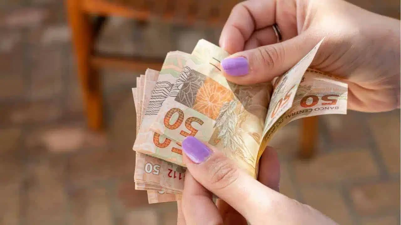 Mãos com esmalte rosa nas unhas contando notas de 50 reais