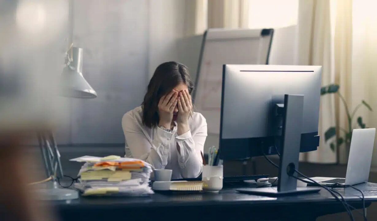 Mulher com mãos tampando o rosto, sentada em um escritório em frente a um computador