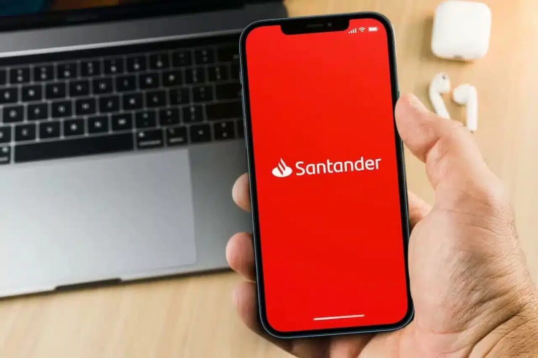 Mão segurando celular que mostra logo do banco Santander. Ao fundo, um notebook.