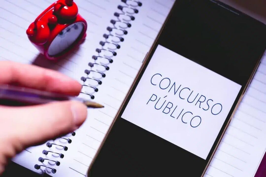Mão segurando caneta. Abaixo, celular com frase "Concurso Público" na tela, sobre um caderno e com uma miniatura de relógio ao lado