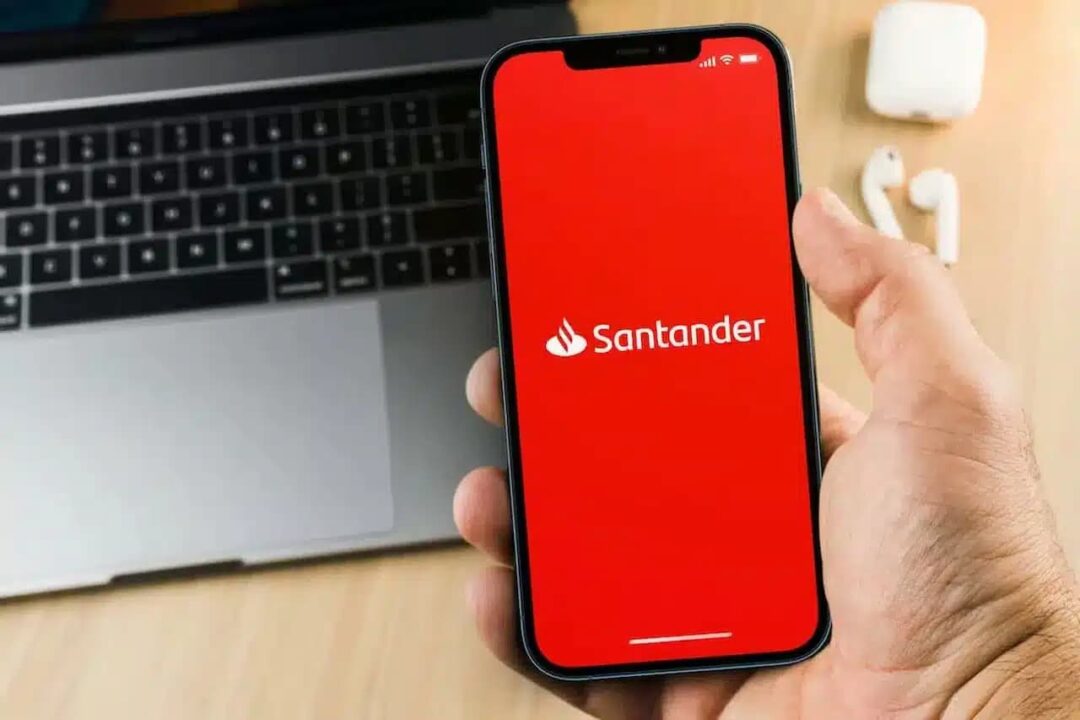 Mão segurando celular que mostra logo do banco Santander. Ao fundo, um notebook