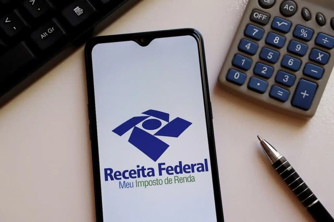 Celular com app do Imposto de Renda aberto na tela, ao lado dele tem uma calculadora e uma caneta