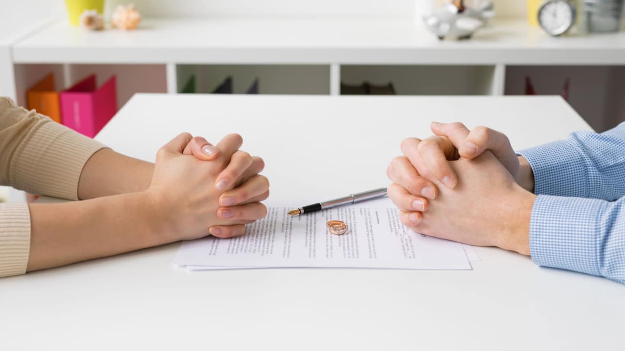 Casal de mãos cruzadas um de frente para o outro enquanto assina papel de divórcio