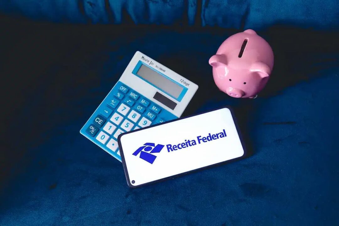 Celular que mostra logo da Receita Federal ao lado de uma calculadora e de um cofrinho em formato de porco