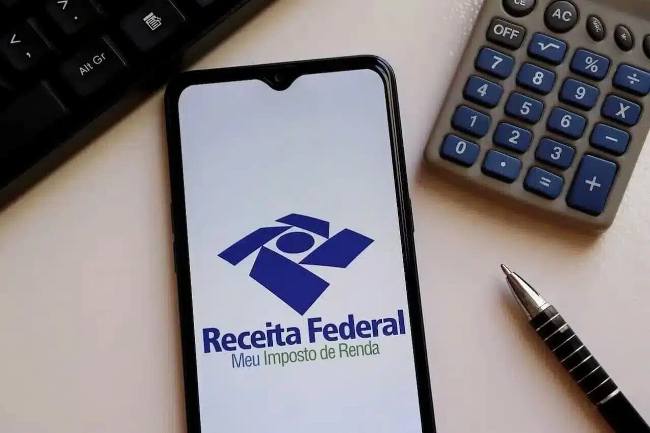 Celular com app do Imposto de Renda aberto na tela, ao lado dele tem uma calculadora e uma caneta IRPF
