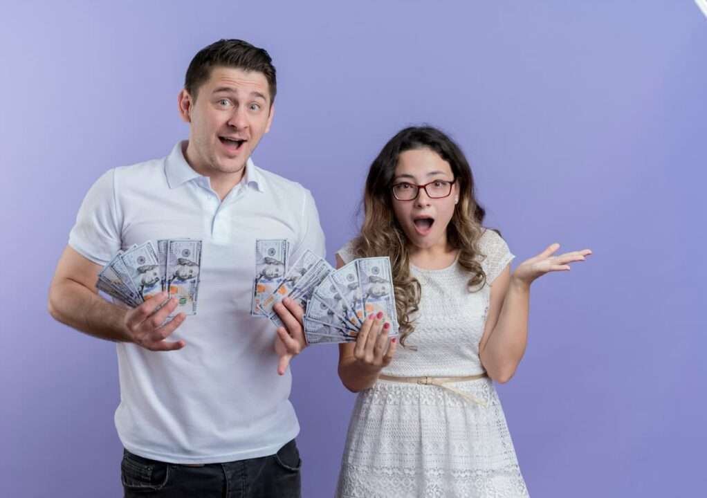 Um homem e uma mulher em pé, ambos com notas de dinheiro na mão e expressões de surpresos