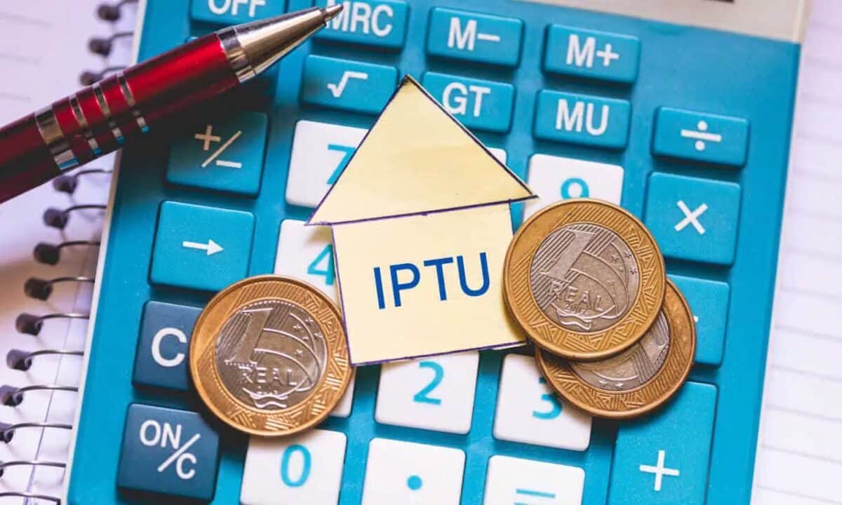 A sigla IPTU dentro de um papel em formato de casa, cercado de moedas, em cima de uma calculadora