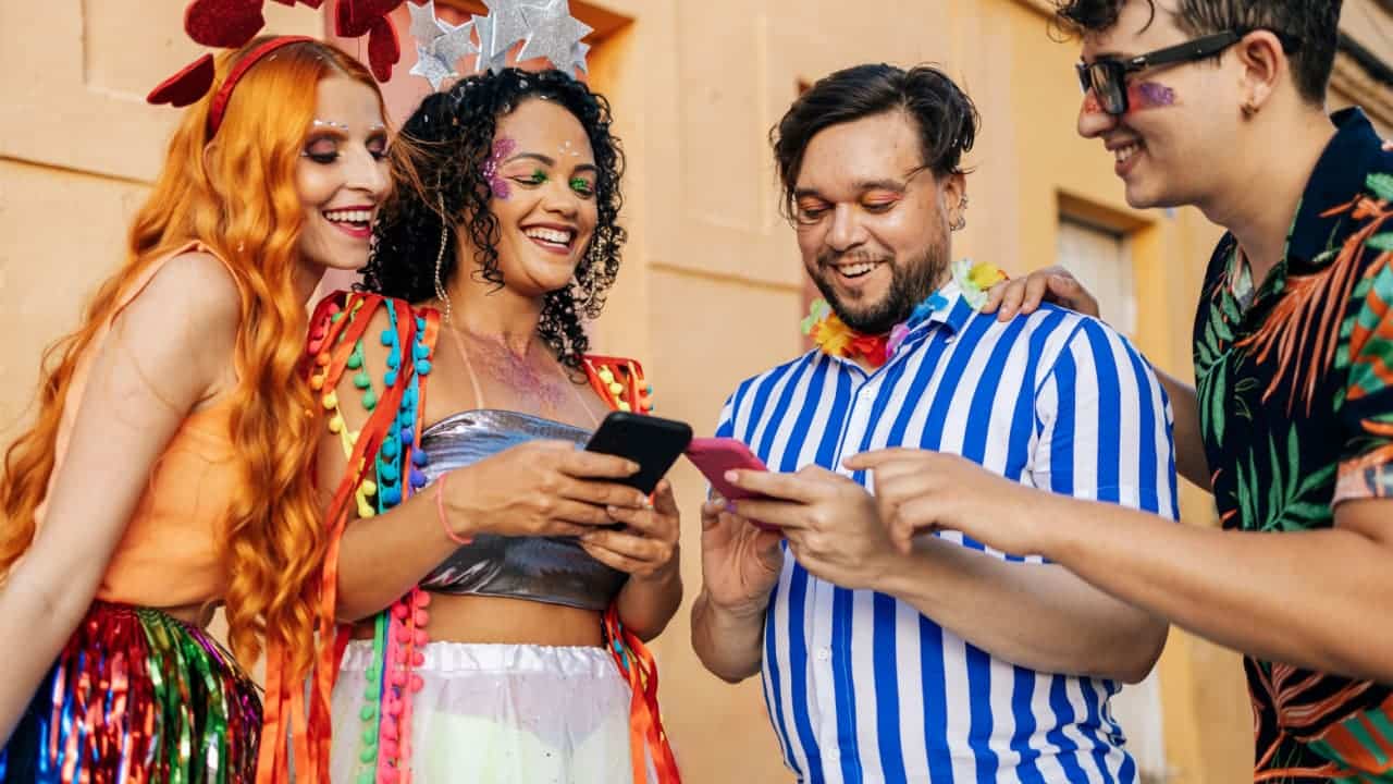 Grupo de quatro amigos em Carnaval na rua, dois deles mexendo no celular enquanto os outros acompanham