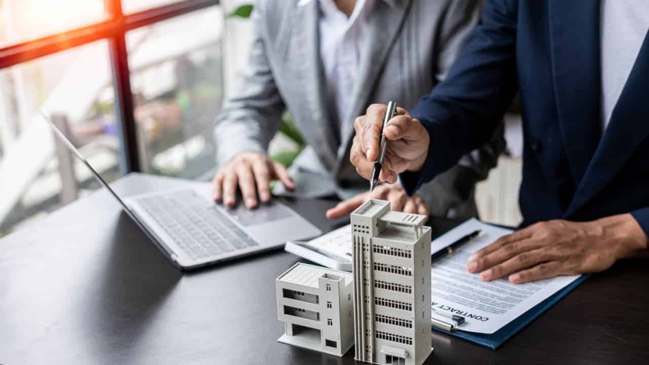 Miniatura de prédios sobre uma mesa, uma pessoa mexendo no computador e outra preenchendo papéis