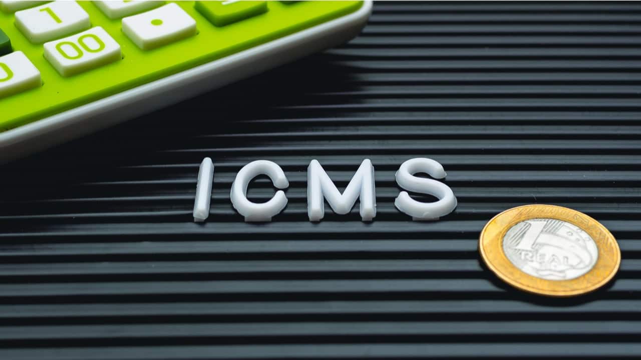 Sigla ICMS escrita em uma placa. Ao lado, há moeda de um real e o pedaço de uma calculadora verde pode ser visto